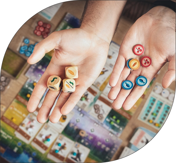 Handen die houten dobbelstenen en kleurrijke speelstukken vasthouden boven een modern strategisch bordspel, waarbij kaarten en onderdelen over de tafel verspreid liggen.