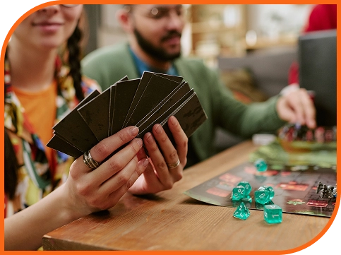 Close-up van spelers die kaarten vasthouden tijdens een bordspelsessie met dobbelstenen, miniaturen en spelonderdelen op een houten tafel.